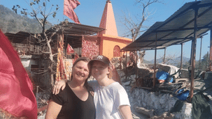 2 people standing together outside an orange temple with a red flag under a clear blue sky.