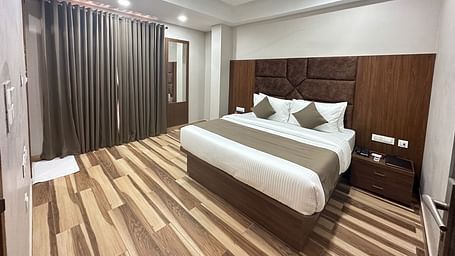A room displays a double bed with white bedding, dark wooden headboard, and wooden flooring - Kankidham Lords Inn