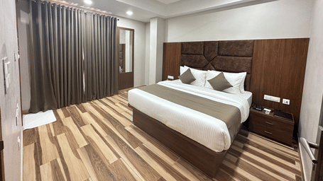A room displays a double bed with white bedding, dark wooden headboard, and wooden flooring - Kankidham Lords Inn