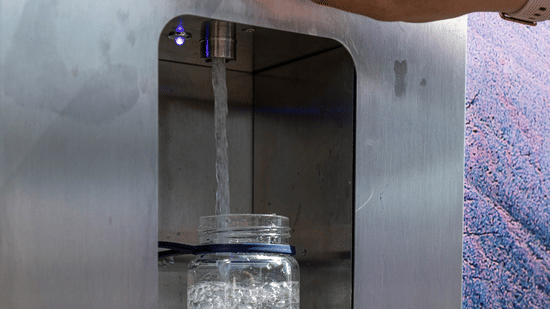 A view of a hand holding a glass bottle beneath the spout of a stainless steel water dispenser, filling the bottle with water.