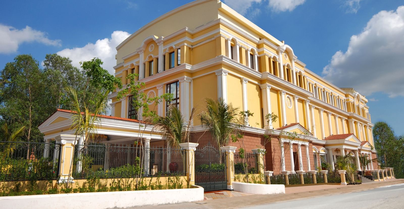 An image of a hotel with an elegant entrance gate, small shrubs lining the pathway, and a lush garden in the foreground