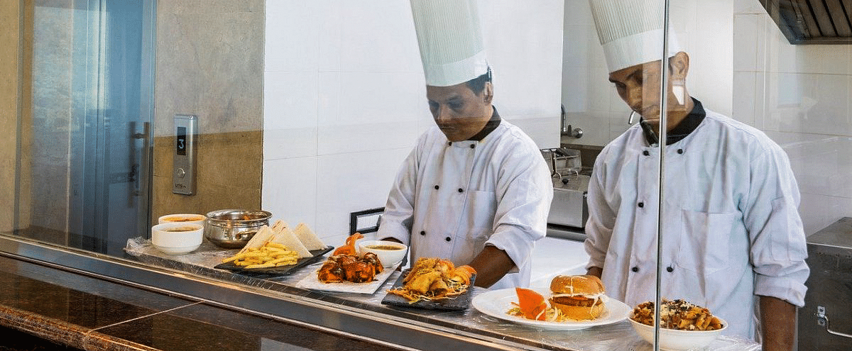 Two chefs preparing dishes in the kitchen as seen from the transparent glass partition at the restaurant at Regenta Inn DLS.