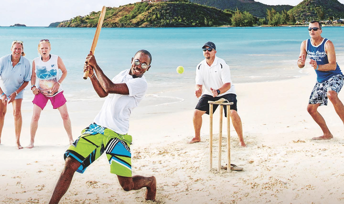 A group of people are playing cricket on a sandy beach with a lush green hill in the background.