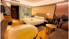 Deluxe King Room at Pride Plaza Ahmedabad showcasing a spacious king size bed, warm wooden flooring, contemporary décor, cosy seating area and a well appointed workspace with soft ambient lighting.