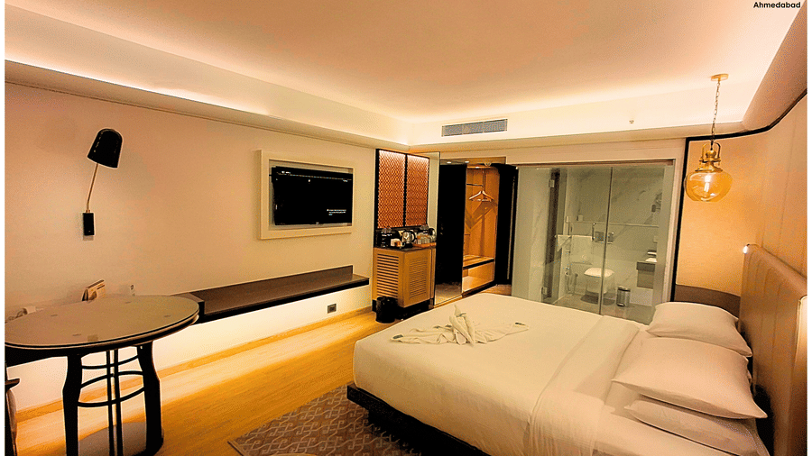 Deluxe King Room at Pride Plaza Ahmedabad displaying a wide angle view of the bedroom with king size bed, television unit, wooden flooring, work desk and elegant ambient lighting.
