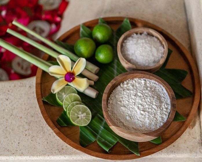 A tray with natural spa elements, including salts, limes, and flowers, on a light-coloured surface.