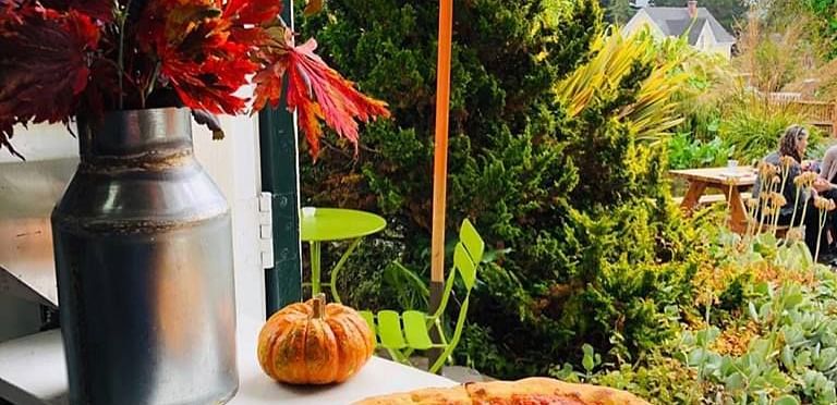 Pizza on a plate by a window, metal vase with foliage, small gourd, umbrella and garden view in background.