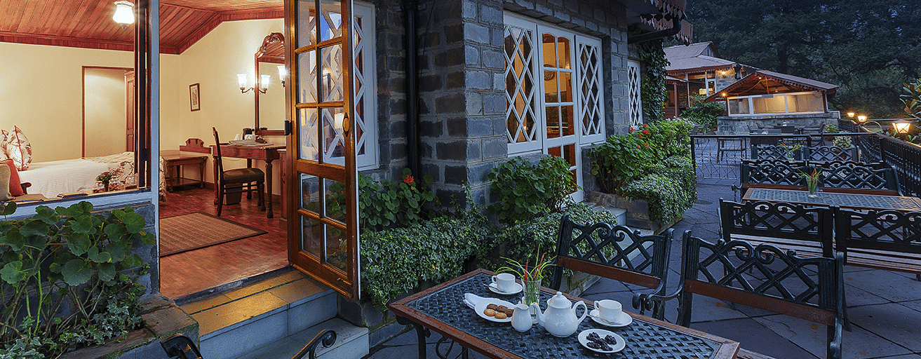 A outer view of the naini retreat, nainital, with a stone exterior and a wooden door leading to a spacious living room with a dining table.,Outside.
