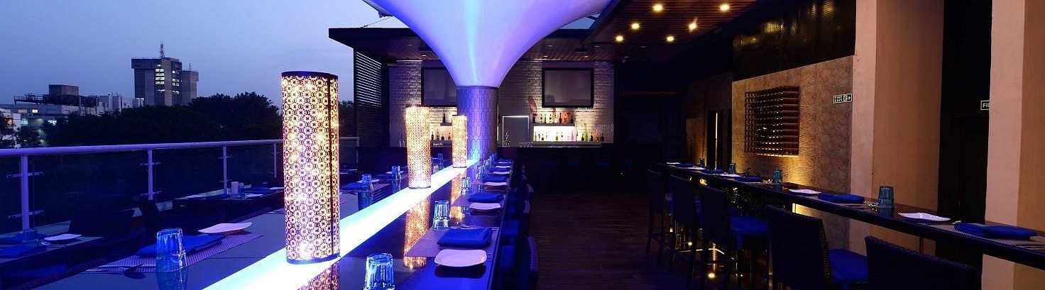 Skylit Rooftop Lounge and Bar at The Cunningham Hotel featuring ambient blue lighting, elegant seating, a modern bar, and a city skyline view at dusk
