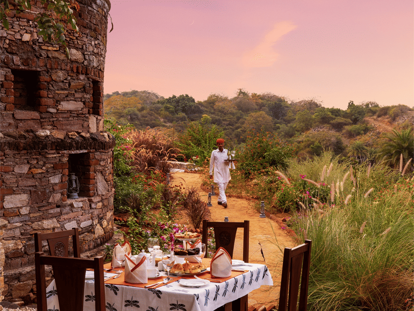 A table for 4 set up next to a stone structure surrounded with greenery and a beautiful sky with pink hues in the background - Chunda Shikar Oudi, Udaipur.