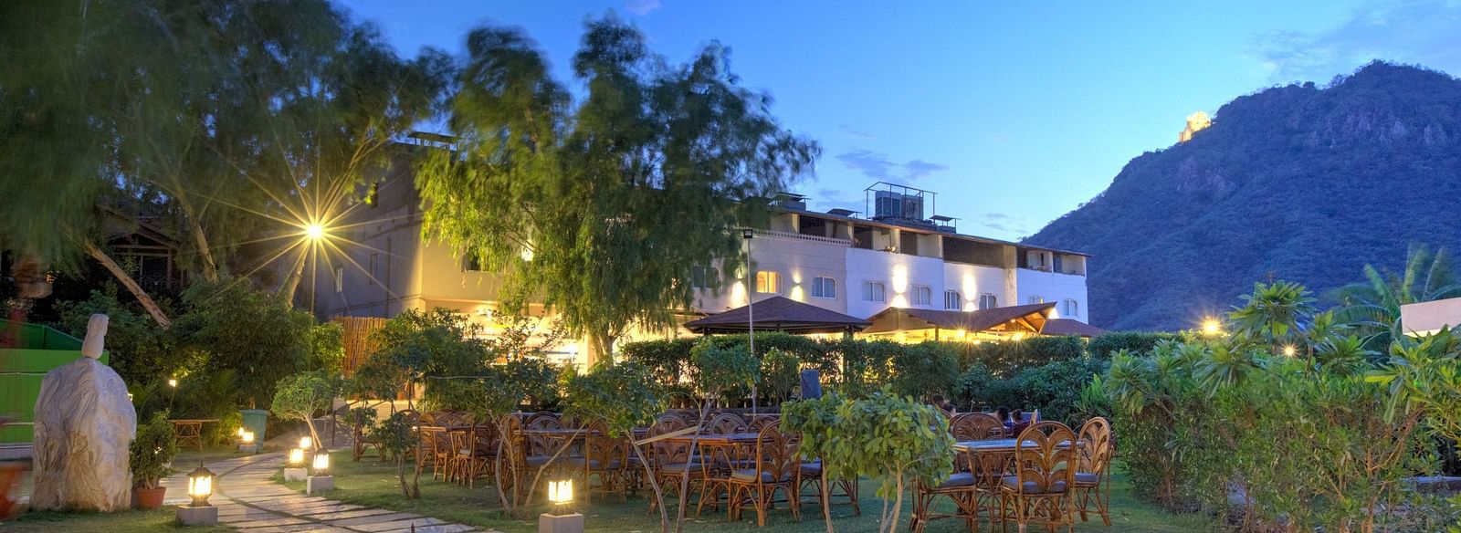 A pathway lit by lanterns leads through a garden at dusk, towards a building with warm lights.- Bamboo Saa Resort & Spa, Udaipur.
