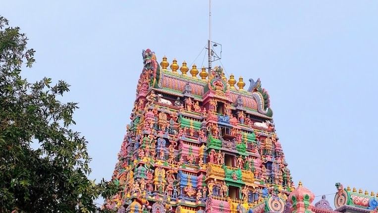 A tall, intricately carved and brightly painted Hindu temple gopuram with colourful statues and decorative details rising against a clear blue sky, partially framed by tree foliage near Chidambara Vilas.