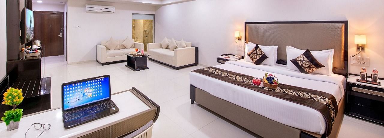 A comfortable hotel suite offers a king size bed, a separate seating area, a functional desk with a laptop, and ample natural light.