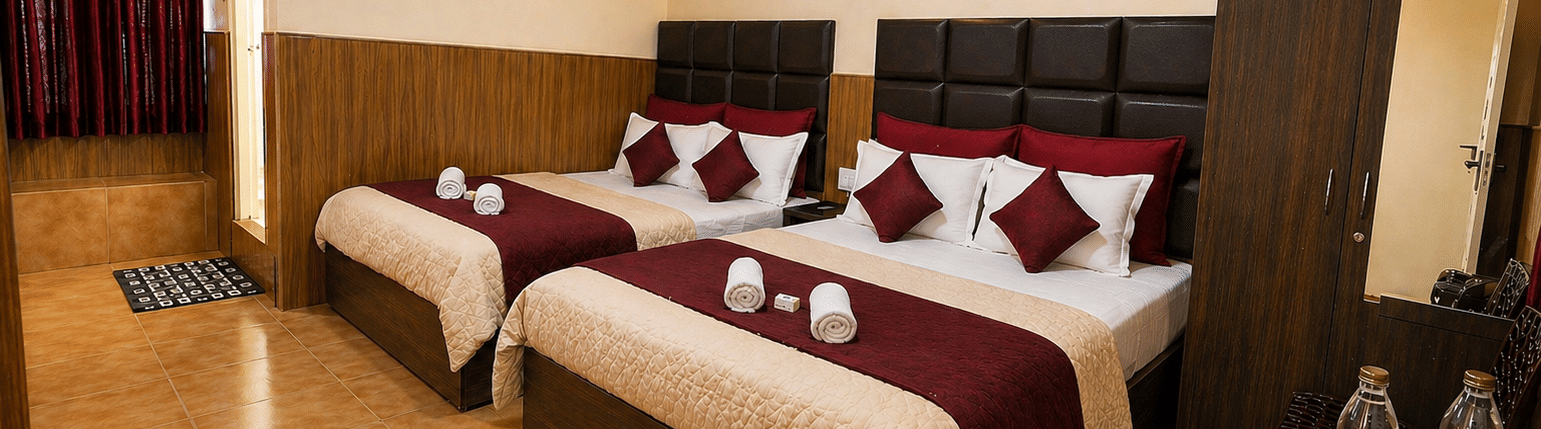 2 large beds arranged side by side with a wardrobe, a wall-mounted fan, and a kettle with water bottles on a table in the Deluxe Quadruple Room at BN Tourist Home, Ooty.