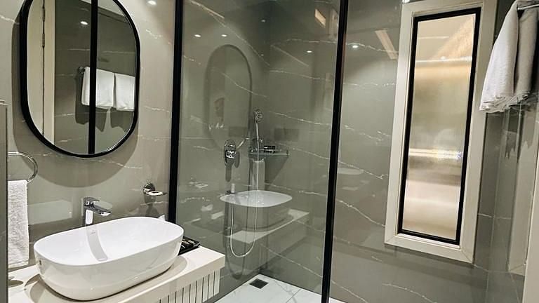 A bathroom area with a western toilet, a shower area, a sink, a mirror and a small window - Five Elements Hotels Goldleaf, Haldwani