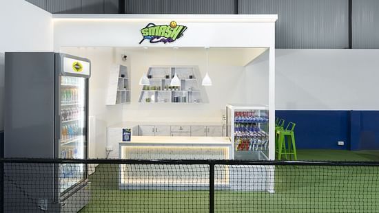 Modern refreshment counter labelled 'Smash Café' with a fridge, seating area, and shelves, located inside the indoor sports facility at Jenneys Residency, Coimbatore.