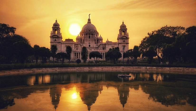 A view of Victoria Memorial, Kolkata, reflected in the water during a golden sunset with trees on either side.