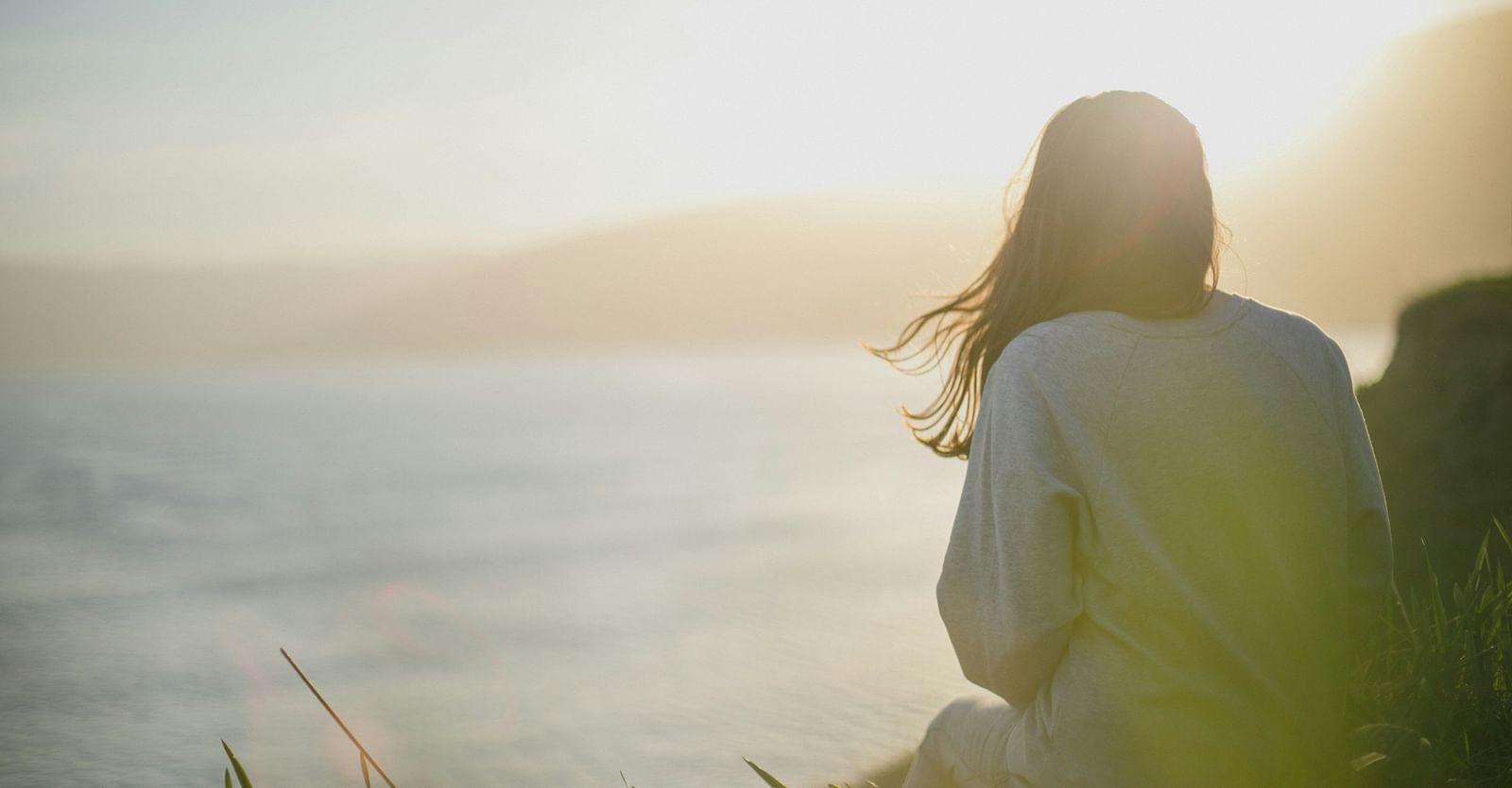 A woman sitting on a cliff while watching the sun near a beach in the distance.