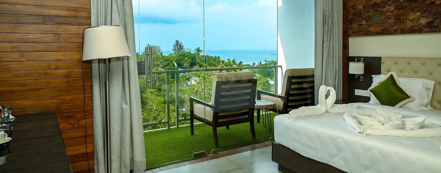 A hotel room interior featuring a large bed, a bedside lamp, and a full-length glass door leading to a balcony with an outdoor seating area, all against a backdrop of wooden wall panelling - Sea Hills Hotels, Sri Vijaya Puram (Port Blair)