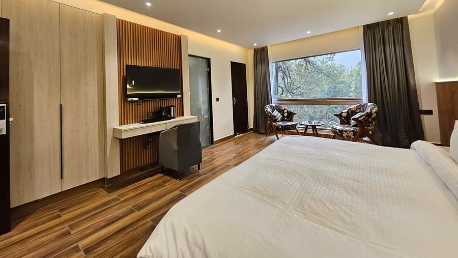 A spacious premium room featuring the bed, flat screen TV, chairs, coffee table, work desk, and an expansive glass window overlooking the view - DLS The Emerald, Nainital