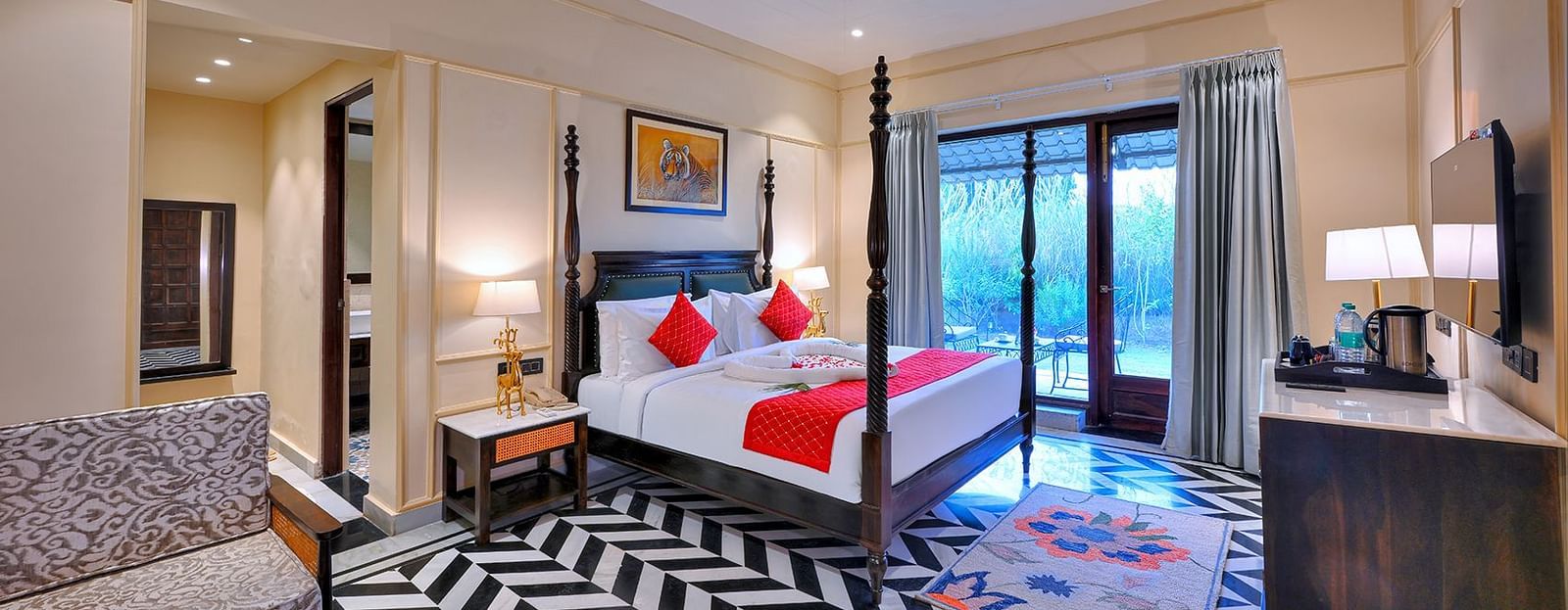 Premium Suite Cottage at Singhvilas Ranthambore features a four-poster bed with red accents, a balcony, a black and white zigzag patterned floor, comfortable seating, a TV, and tiger-themed artwork.