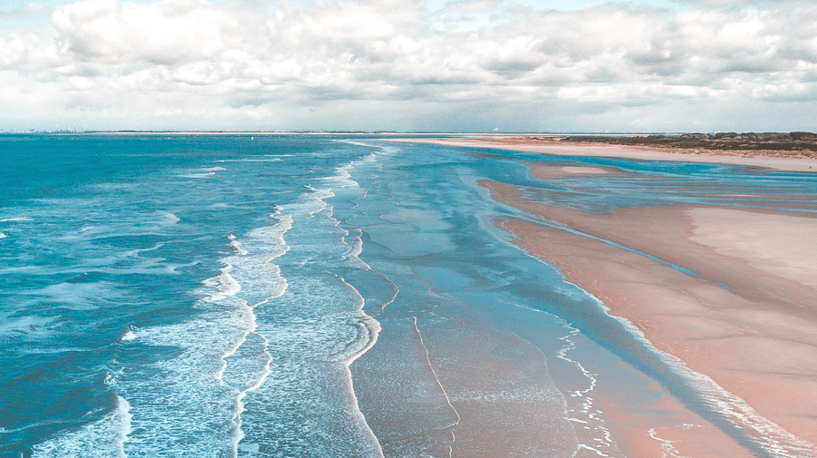 An aerial view of a sandy beach with gentle waves rolling onto the shore, under a bright blue sky with scattered clouds.