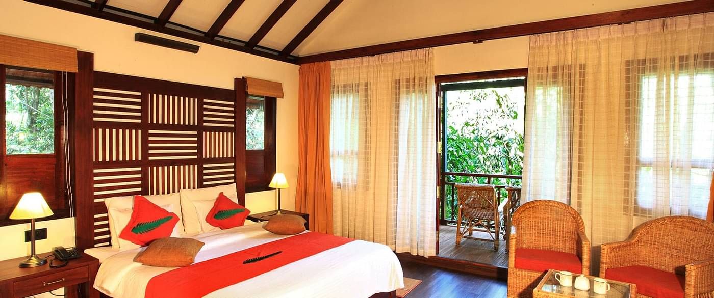 A bed placed in a room with two chairs and table, and a balcony that offers forest views | Vythiri Resort, Wayanad