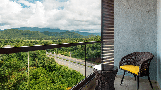  A chair and coffee table on the balcony of one of the rooms offer a view of lush trees and mountains under a cloudy sky at Cloud9 Sarovar Premiere, Palolem Goa
