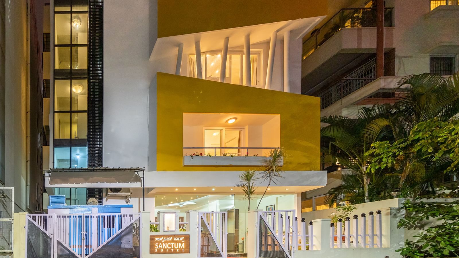 A facade of Sanctum Suites Domlur, Bangalore with warm lighting and view of balconies shot at night.