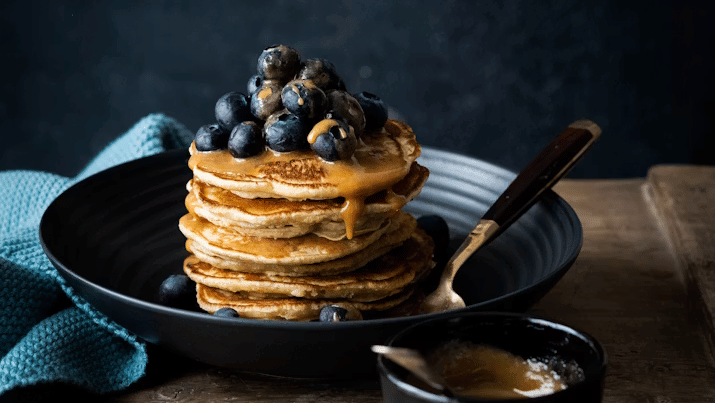 Stack of pancakes topped with blueberries and dripping maple syrup on a black plate, served with a spoon and a small black bowl of syrup.