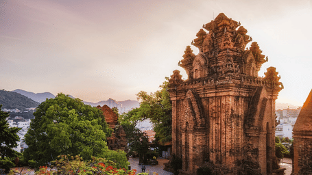 An overview of Po Nagar Temple, a monumental structure, near some bushes with a waterbody in the background, as seen during sunset.