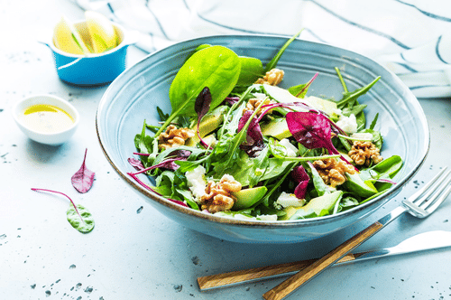 A fresh green salad in a blue bowl with walnuts, avocado, and red cabbage, with dressing and lemons