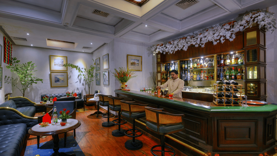 A bar with a man standing near the counter, bottles placed on shelves, sofas, and tables placed infront of the counter.