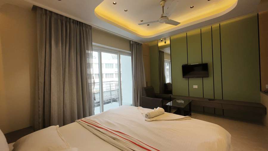 Deluxe Classic Varanda Room featuring a large bed with white linens, grey curtains covering a glass door to a balcony, a ceiling fan, and a wall-mounted television on wall at Hotel Seagull Digha