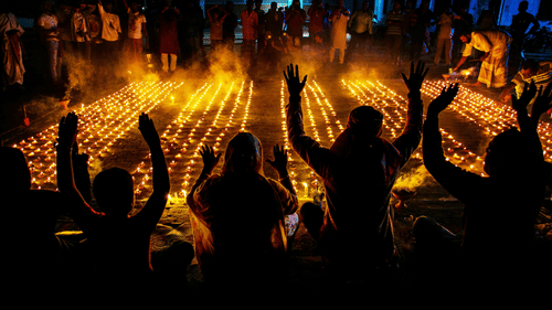 A group of people with raised arms standing around rows of brightly-lit oil lamps during the night.