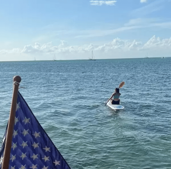 A person paddles a stand-up paddle board in the ocean, framed by the corner of a large American flag.