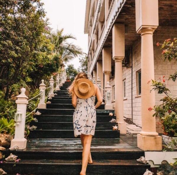 A woman with blonde hair hiding her face with a hat as she poses for a photograph standing on the elegant staircase surrounded with plants and flowery bushes.