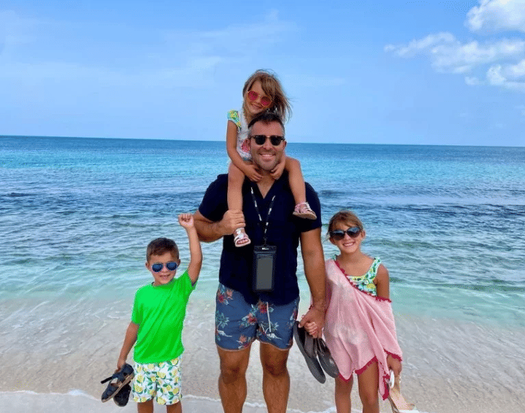 A family on a sunny beach, with a man carrying a child on his shoulders, facing the ocean