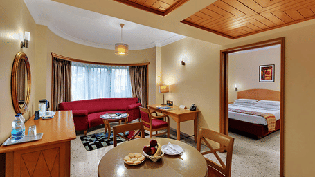 A hotel suite living area with a red sofa, dining table, and a doorway leading into a bedroom | Trance Oakwood Hotel, Pune