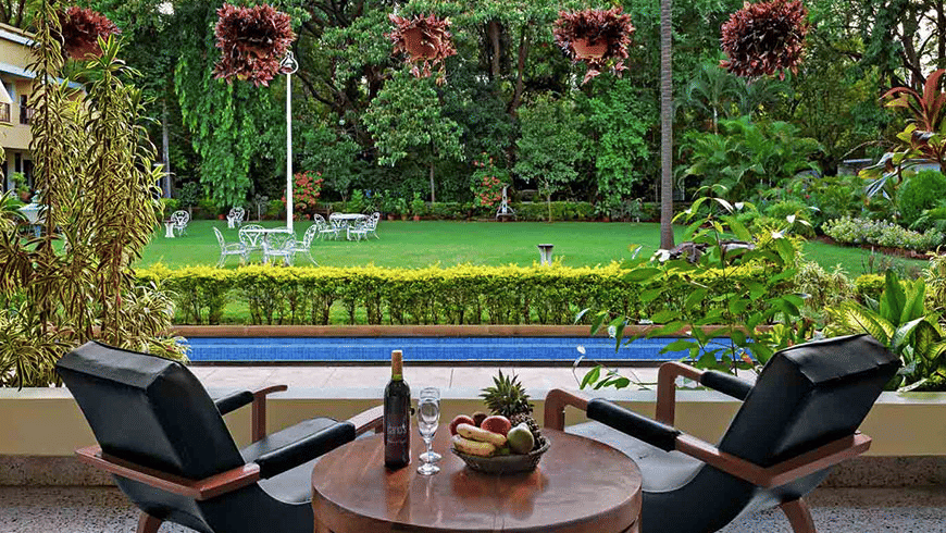 2 black lounge chairs facing a small round table with a bottle and fruit, overlooking a pond and garden at Trance Sunderban Resort.