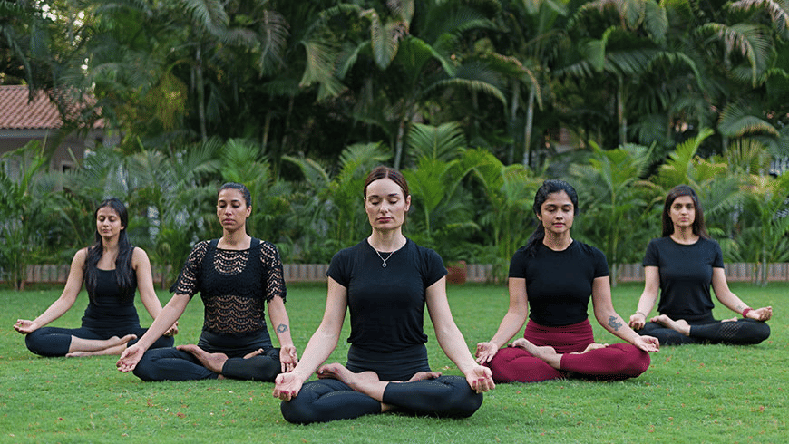 A group of 3 women sitting in a row on a green lawn at Trance Sunderban Resort, performing yoga in front of dense tropical trees.