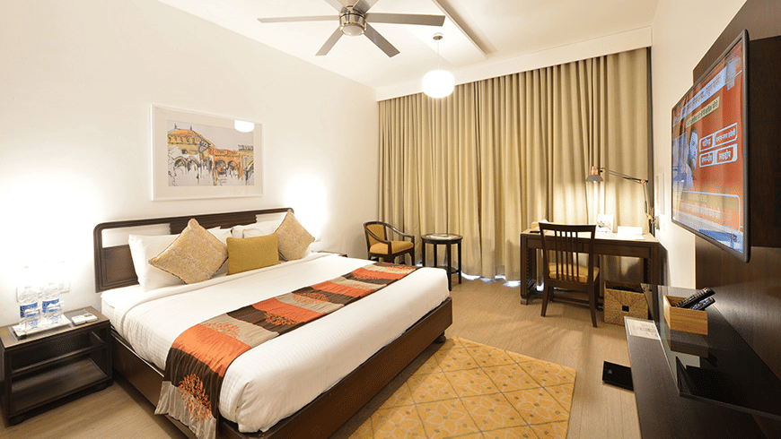 A well-lit Studio Room at Trance Sunderban Resort featuring a bed with orange accents, a desk, and a television.