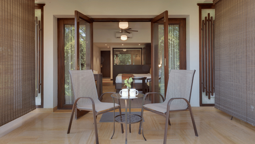 2 grey mesh chairs and a small glass table set on a tiled balcony at Trance Sunderban Resort, with open doors leading into a room.