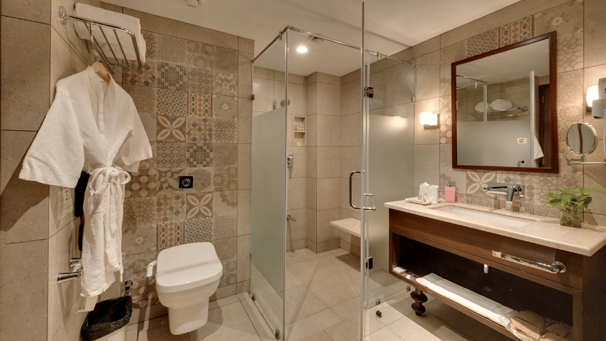A modern bathroom at Trance Sunderban Resort with patterned wall tiles, a glass shower, a large mirror, and a bathrobe.
