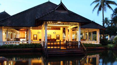 A traditional, open-sided tropical pavilion with a dark thatched roof, warmly lit from within, overlooking a calm body of water at dusk.