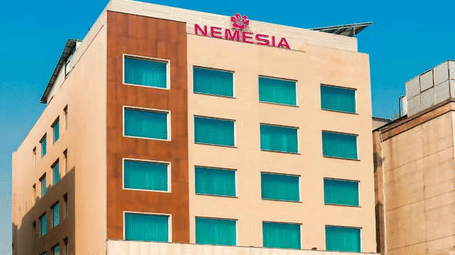 Daytime view of the Nemesia hotel building facade with teal-tinted windows, wooden accents, and palm trees in front.