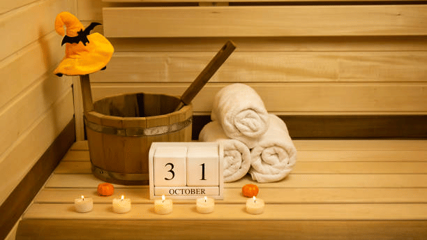A wooden sauna bench with a bucket, rolled white towels, tea lights, mini pumpkins and a witch hat beside a 31 October calendar block