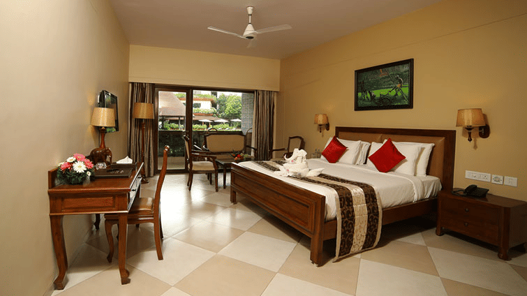 Mystica Room at Uday Suites featuring a plush bed, desk, balcony with chairs and a ceiling fan under warm lighting