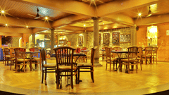 The warmly lit Lounge of Uday Samudra Leisure Beach Hotel features wooden tables and chairs arranged on a raised platform with pillars.