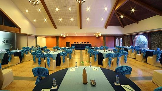 A spacious Vizhinjam Hall at Uday Samudra Leisure Beach Hotel, Kovalam, is prepared for an event with round tables and blue-draped chairs.